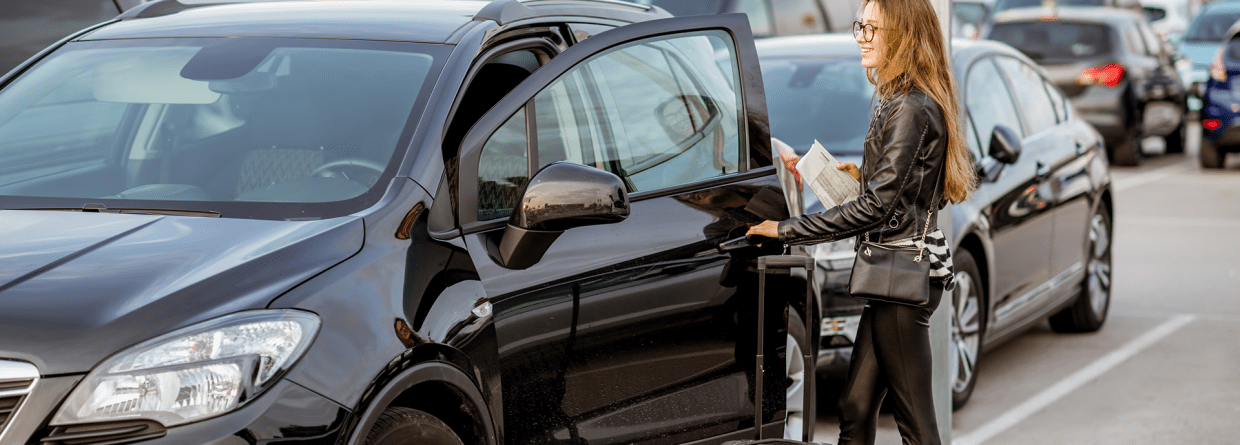 Jonge, vrolijke vrouw met een koffer en papieren in haar hand op een terrein waar auto's verhuurd worden